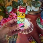 A hand with pink nails holds a colorful bundle of dried herbs tied with white string, featuring red, yellow, green, and blue sections. The background is a styled tabletop with flowers, a shell, and a warm orange surface, with a 'SMUDGE SPIRITUAL BOUTIQUE ENGLAND' logo at the bottom left.