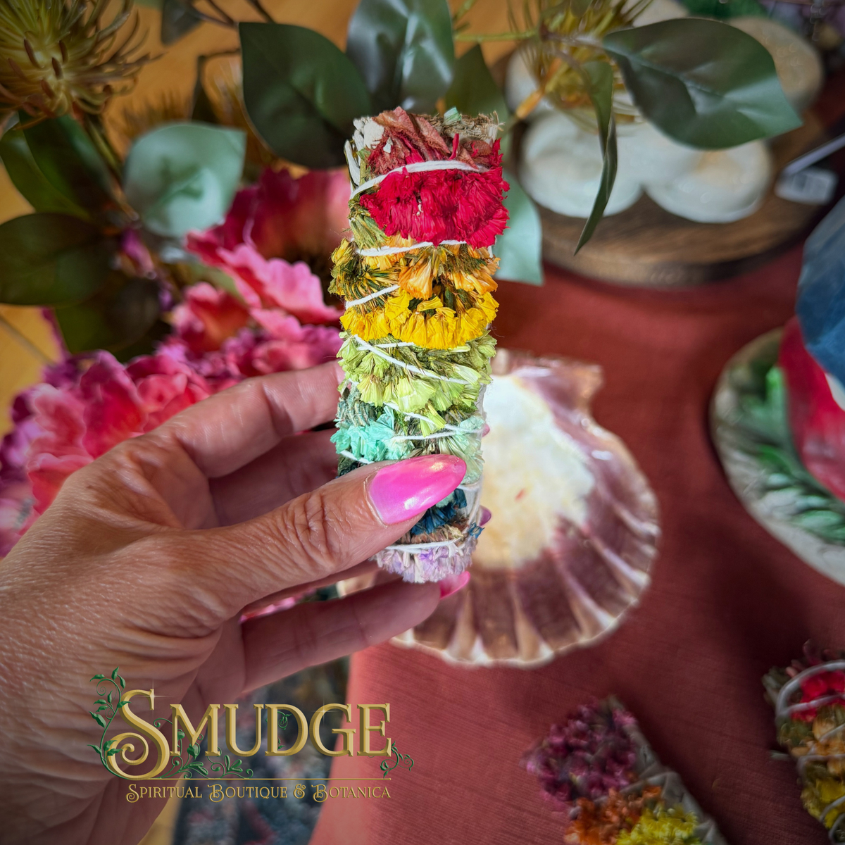 A hand with pink nails holds a colorful bundle of dried herbs tied with white string, featuring red, yellow, green, and blue sections. The background is a styled tabletop with flowers, a shell, and a warm orange surface, with a 'SMUDGE SPIRITUAL BOUTIQUE ENGLAND' logo at the bottom left.