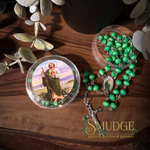 A green beaded rosary with a silver-toned crucifix and a round clear case containing matching green beads, arranged on a wooden surface with decorative leaves. The case lid shows a printed religious illustration.