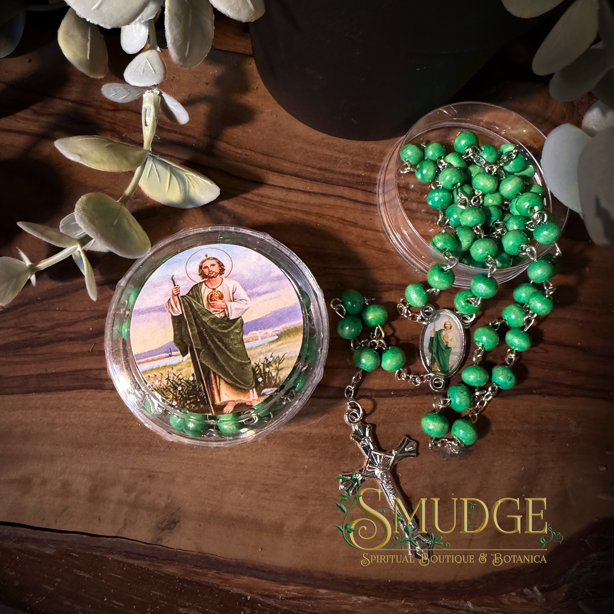 A green beaded rosary with a silver-toned crucifix and a round clear case containing matching green beads, arranged on a wooden surface with decorative leaves. The case lid shows a printed religious illustration.