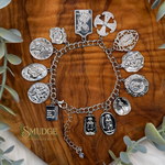 A silver-tone charm bracelet featuring multiple religious medallion pendants, displayed on a polished olive wood background with eucalyptus leaves. The brand name 'SMUDGE' is visible in the bottom-left corner.