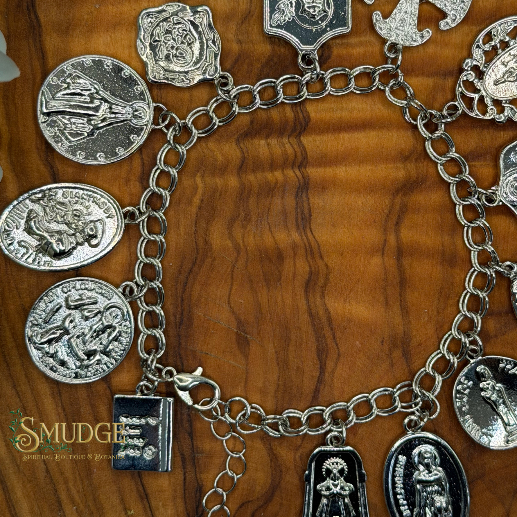 A silver-tone charm bracelet with multiple religious and saint-themed pendants, displayed on a polished wooden surface. The bracelet features various charms including oval saint medallions, shield emblems, and a small book charm.