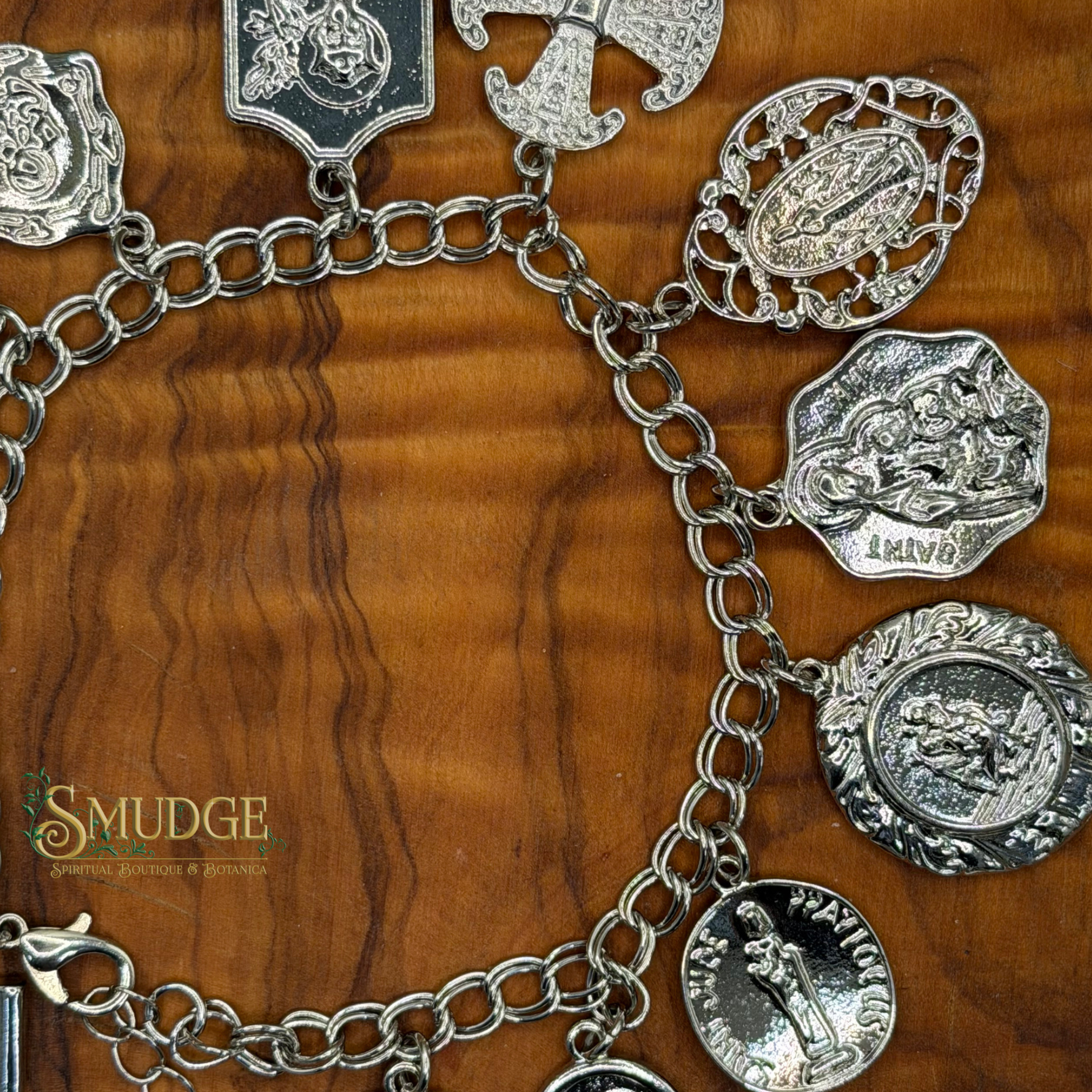 A silver-tone charm bracelet with multiple religious medal and coin-shaped charms, displayed on a warm brown wood grain background. The brand name 'Smudge' is visible as a watermark in the lower-left corner.