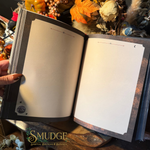 An open journal or notebook with blank cream-colored pages, held by a hand. The cover and inner margins have a dark, gothic-style design with celestial illustrations like stars and a crescent moon.