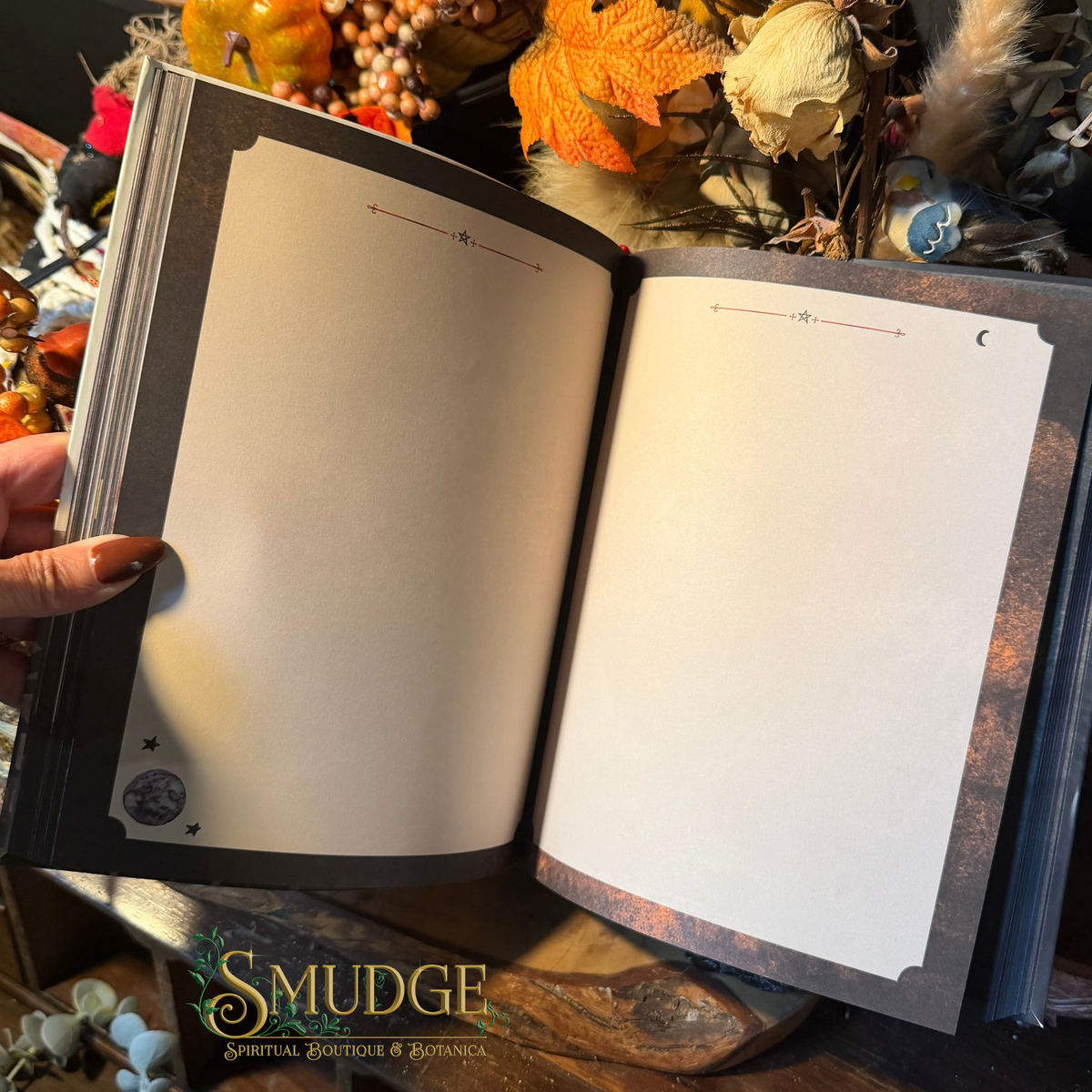 An open journal or notebook with blank cream-colored pages, held by a hand. The cover and inner margins have a dark, gothic-style design with celestial illustrations like stars and a crescent moon.