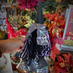 A hand holds a small witch-themed decorative figure with a black pointed hat, black fringe, and iridescent purple spiderweb-patterned fabric, set against a busy floral and seasonal display with a visible 'Smudge' watermark.