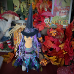 A witch-themed plush doll with a black hat, purple and blue iridescent dress, and long purple string hair, displayed among autumn leaves, mushrooms, and a jar in a decorative indoor setting. A sign reading 'Smudge' is visible in the background.