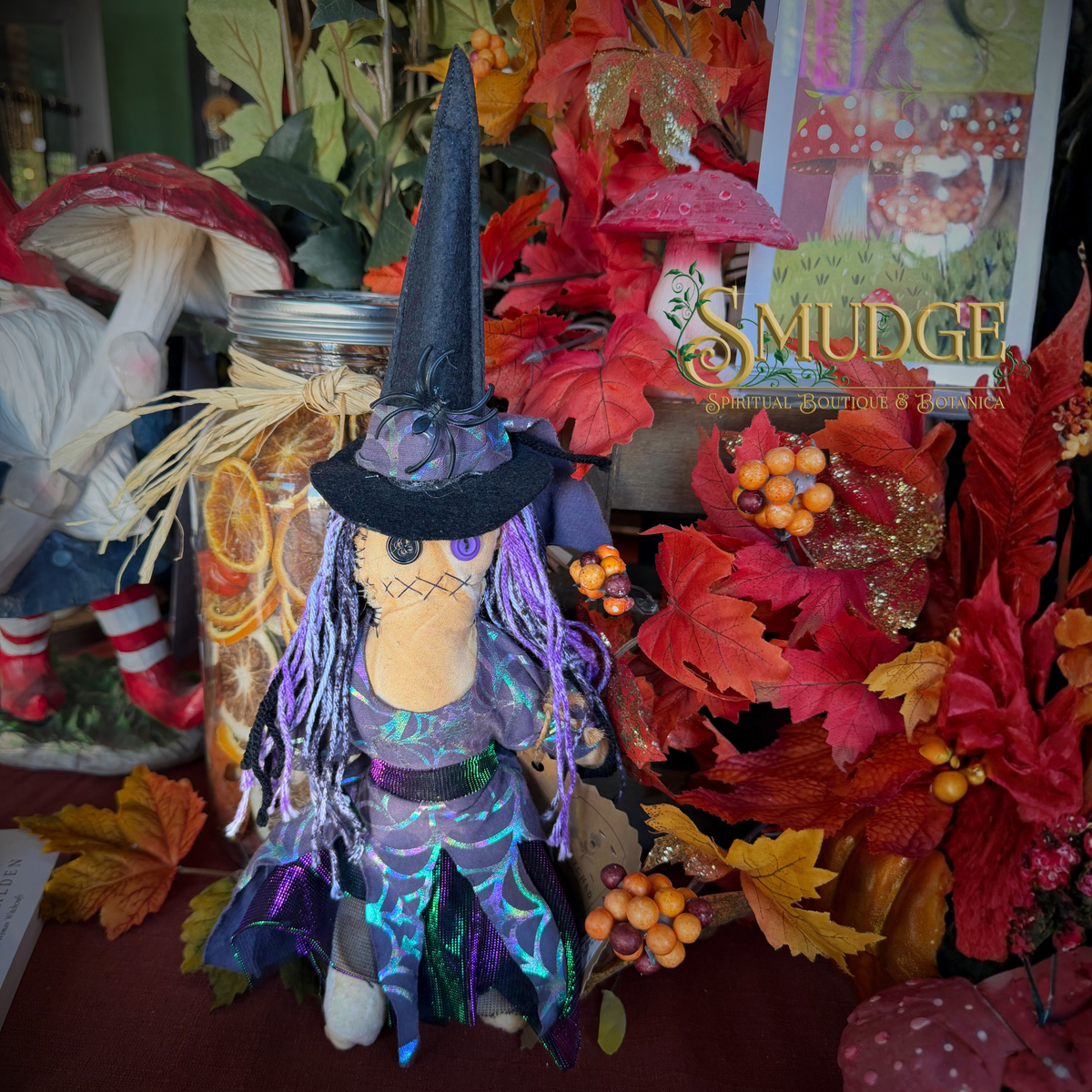 A witch-themed plush doll with a black hat, purple and blue iridescent dress, and long purple string hair, displayed among autumn leaves, mushrooms, and a jar in a decorative indoor setting. A sign reading 'Smudge' is visible in the background.