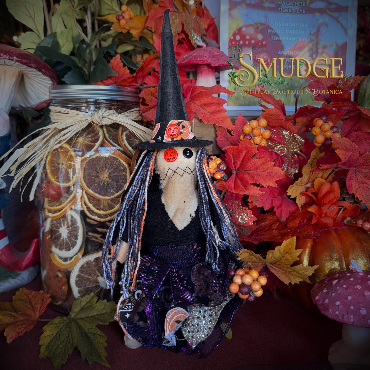 A handmade witch doll with a black pointed hat, purple dress, and orange nose, styled in a cozy autumn display with dried orange slices in a jar, red leaves, and mushrooms. A sign reading 'Smudge' is visible in the background.