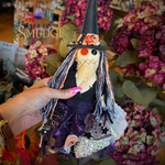 A handmade witch scarecrow doll with a black hat, orange pumpkin face, and purple dress, held by a hand in a floral shop. The brand name 'Smudge' is visible as an overlay.