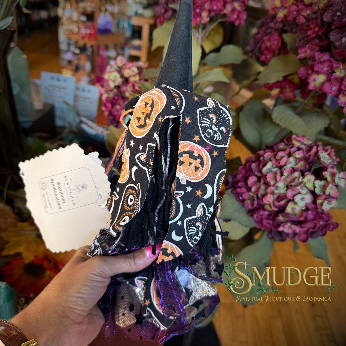 A hand holds a black witch hat decorated with a Halloween pattern of orange pumpkins, black cats, and stars, featuring purple fringe and tinsel. The background shows a shop interior with purple hydrangeas and a 'SMUDGE SPIRITUAL BOUTIQUE & BOTANICA' logo overlay.