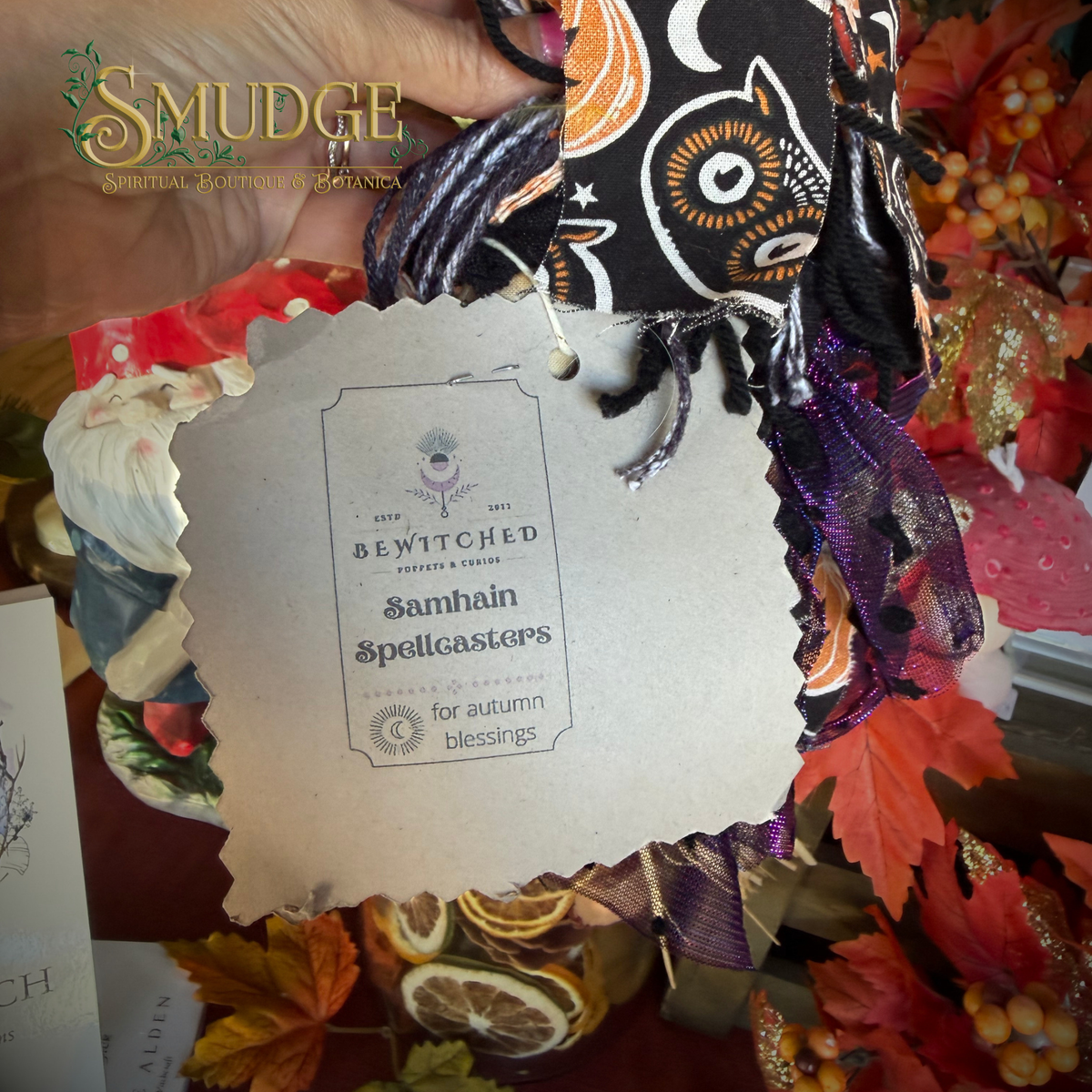 A hand holds a bundle of autumn-themed fabric scraps with a paper tag reading 'Samhain Spellcasters' from Swicthed. The fabrics include black with orange and white owl patterns, purple and black striped ribbon, and other seasonal prints, surrounded by fall leaves, dried citrus, and berries.