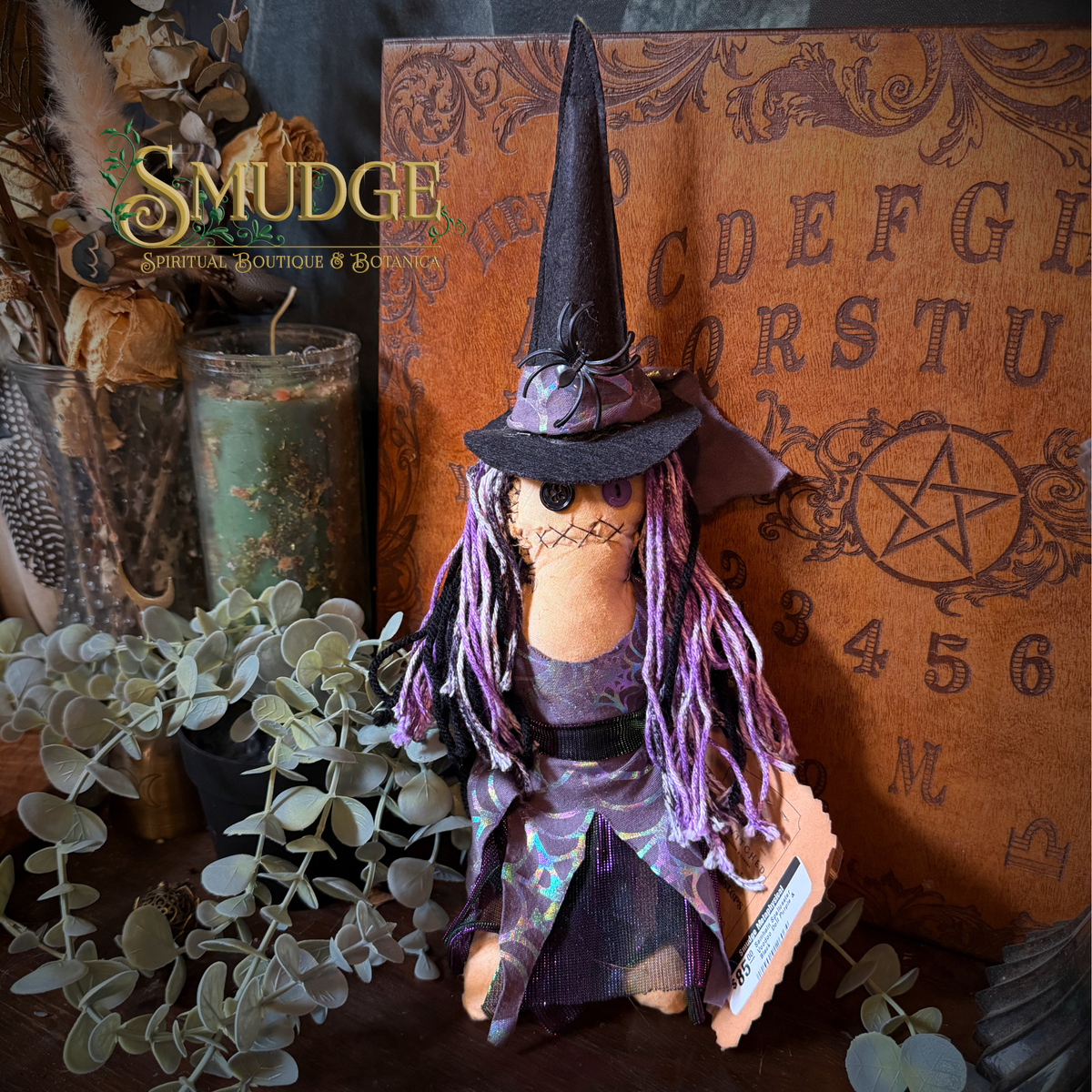 A small witch doll with a black pointed hat, purple yarn hair, and a dark dress stands in a styled tabletop scene with eucalyptus leaves, candles, and a brown almanac in the background. The brand name 'SMUDGE' appears in the upper left.