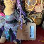 A close-up of a sewn cloth Halloween decoration featuring a stitched face, purple yarn hair, and a shimmering purple dress with star patterns. A paper tag is attached, and dried orange slices and a jar labeled 'Smudge' are visible in the background.