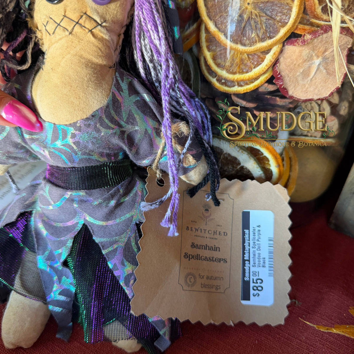 A close-up of a sewn cloth Halloween decoration featuring a stitched face, purple yarn hair, and a shimmering purple dress with star patterns. A paper tag is attached, and dried orange slices and a jar labeled 'Smudge' are visible in the background.