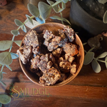 A rustic brown ceramic bowl filled with dried, sandalwood resin incense pieces, styled on a dark wooden table with green eucalyptus leaves and a dark pot in the background. The brand name 'Smudge spiritual Boutique & Botanica' is watermarked at the bottom left.