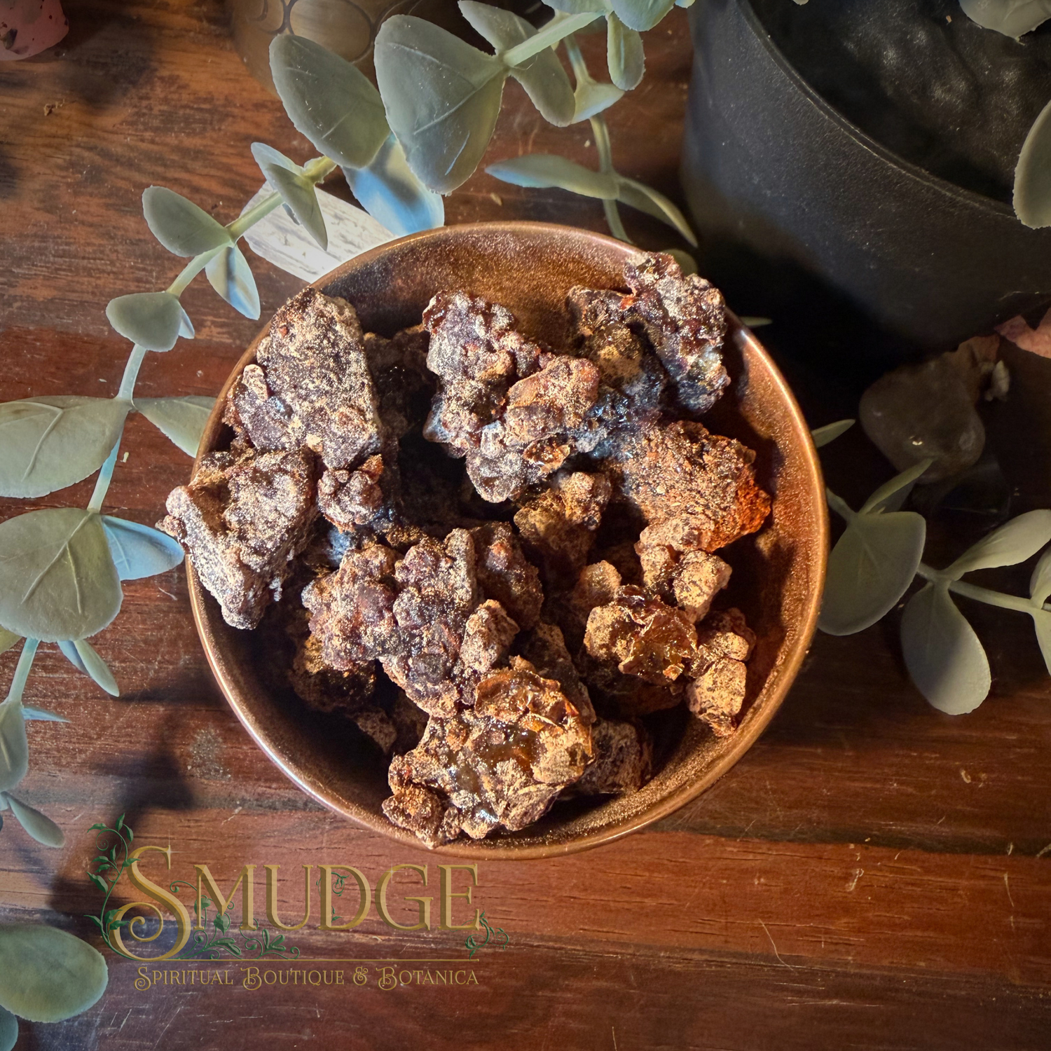 A rustic brown ceramic bowl filled with dried, sandalwood resin incense pieces, styled on a dark wooden table with green eucalyptus leaves and a dark pot in the background. The brand name 'Smudge spiritual Boutique & Botanica' is watermarked at the bottom left.