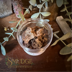 A small clear plastic jar filled with brown sandalwood resin incense, placed on a dark wooden surface with decorative leaves, crystals, and a visible 'Smudge Spiritual Boutique & Botanika' logo at the bottom.