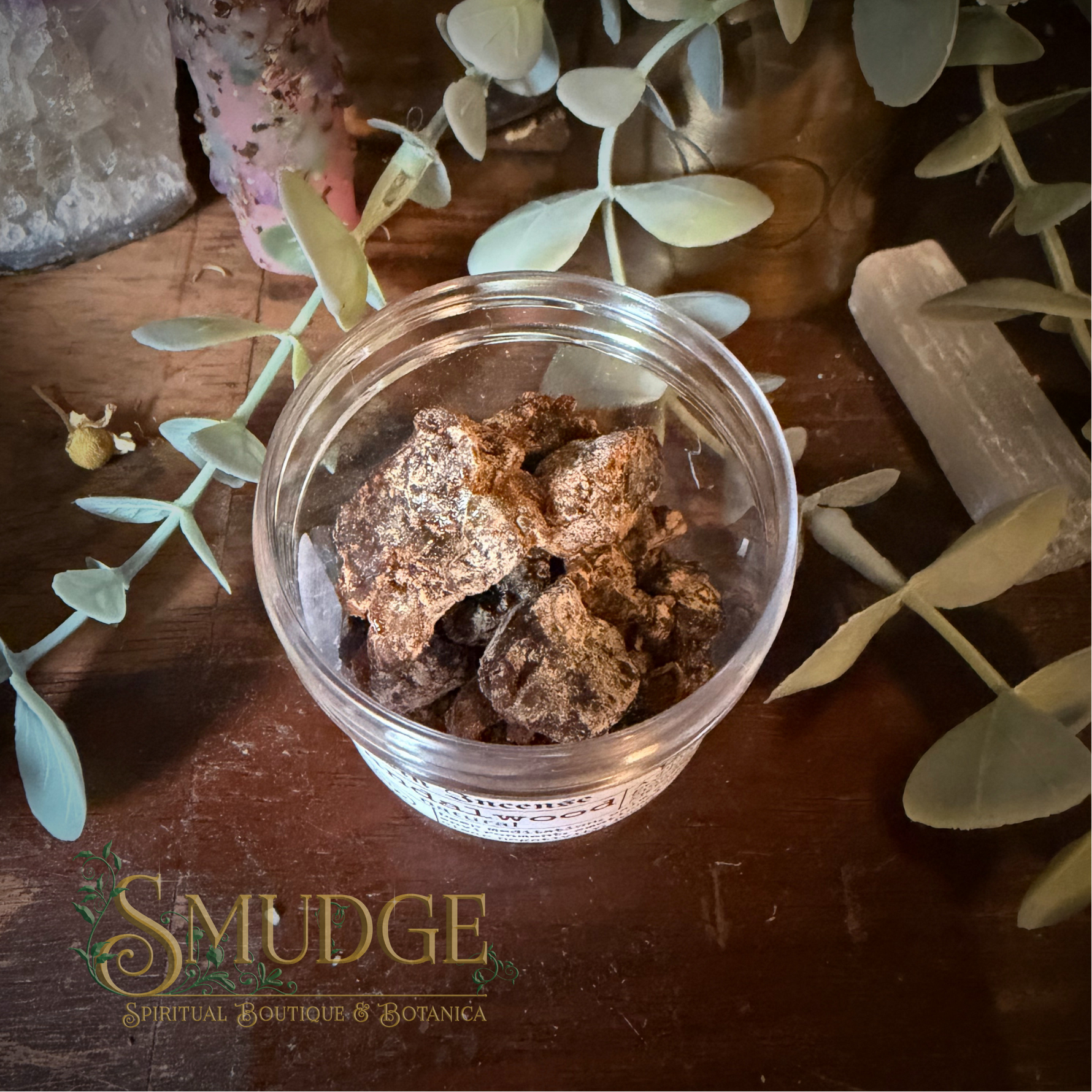 A small clear plastic jar filled with brown sandalwood resin incense, placed on a dark wooden surface with decorative leaves, crystals, and a visible 'Smudge Spiritual Boutique & Botanika' logo at the bottom.