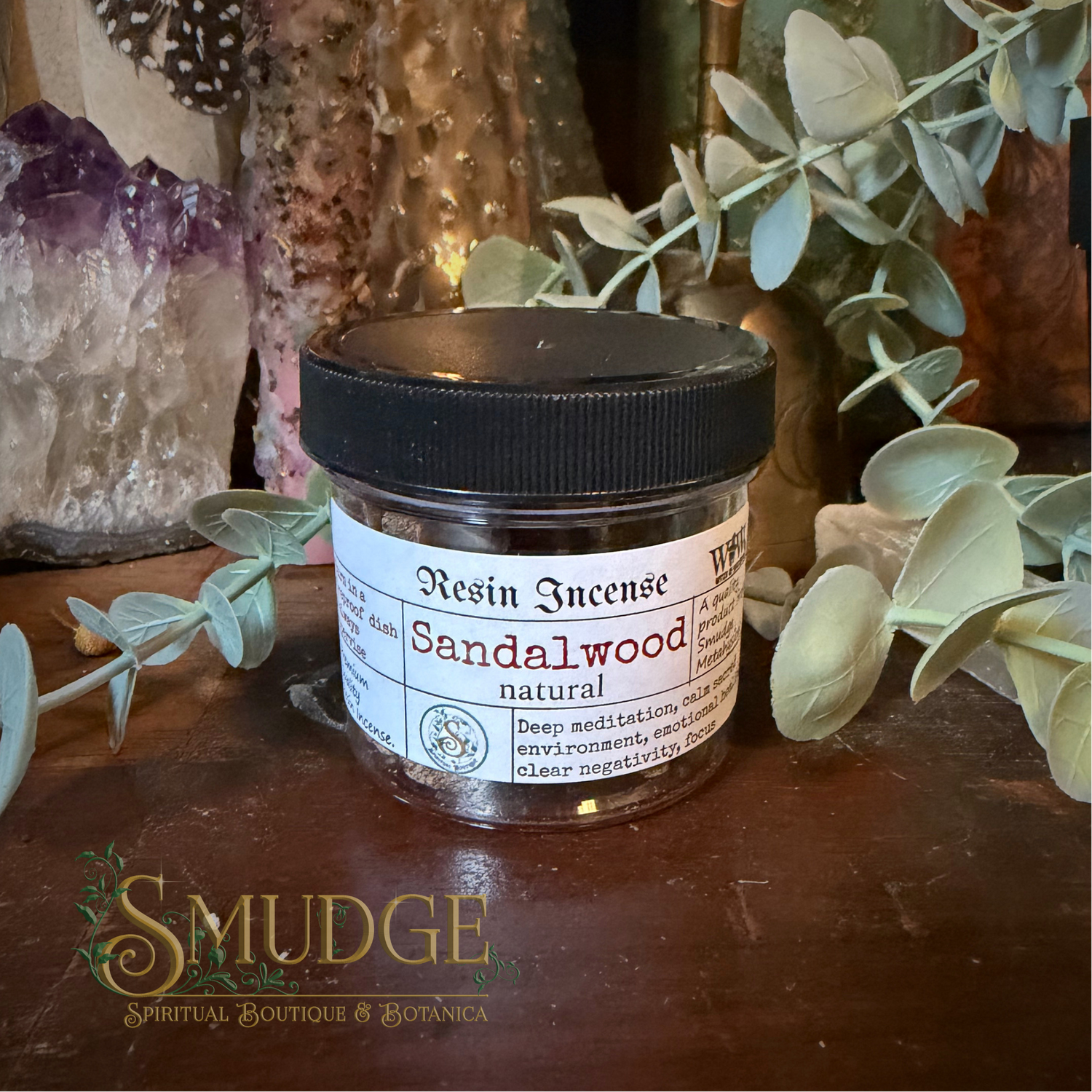 A small clear plastic jar of resin incense with a black lid and a white label reading 'Sandalwood natural,' placed on a wooden surface with decorative crystals, eucalyptus branches, and a Smudge Boutique logo at the bottom.