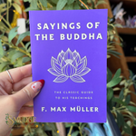 A hand holds a purple book titled 'Sayings of the Buddha' by F. Max Muller, featuring a white lotus illustration on the cover, with a blurred background of green plants.