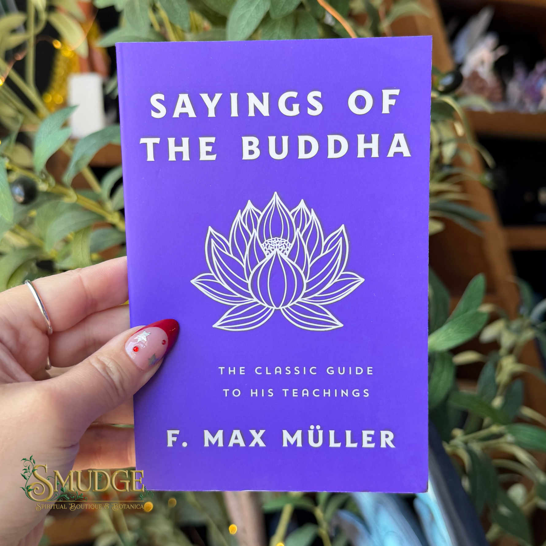 A hand holds a purple book titled 'Sayings of the Buddha' by F. Max Muller, featuring a white lotus illustration on the cover, with a blurred background of green plants.