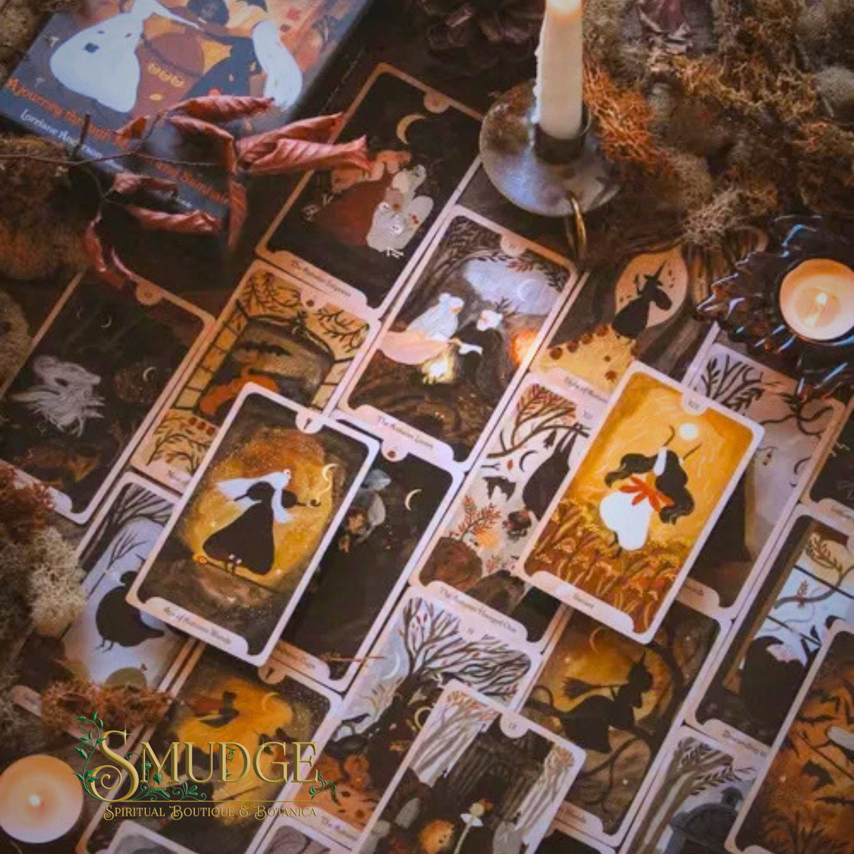 A styled flat-lay of illustrated tarot cards spread across a dark surface, surrounded by candles, dried leaves, moss, and autumn-themed props. The cards feature dark fantasy and gothic artwork in black, gold, and brown tones.
