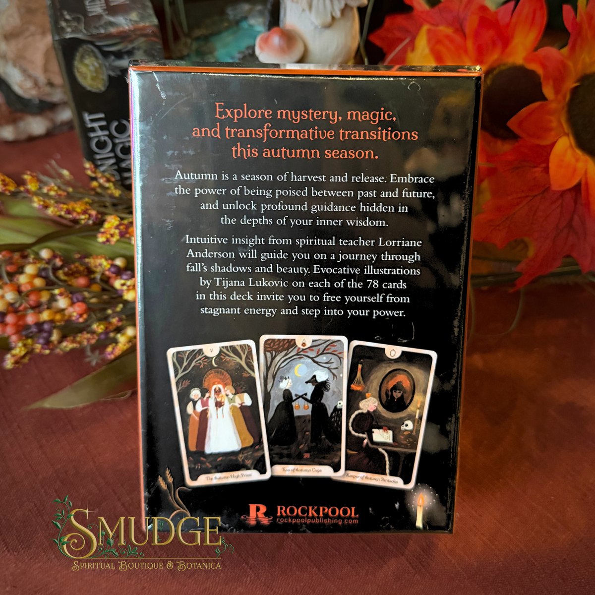 The back of the Rockpool Oracle Cards deck box is shown on a wooden surface with autumn-themed flowers in the background. The box is black with orange text and illustrations of three oracle cards at the bottom.