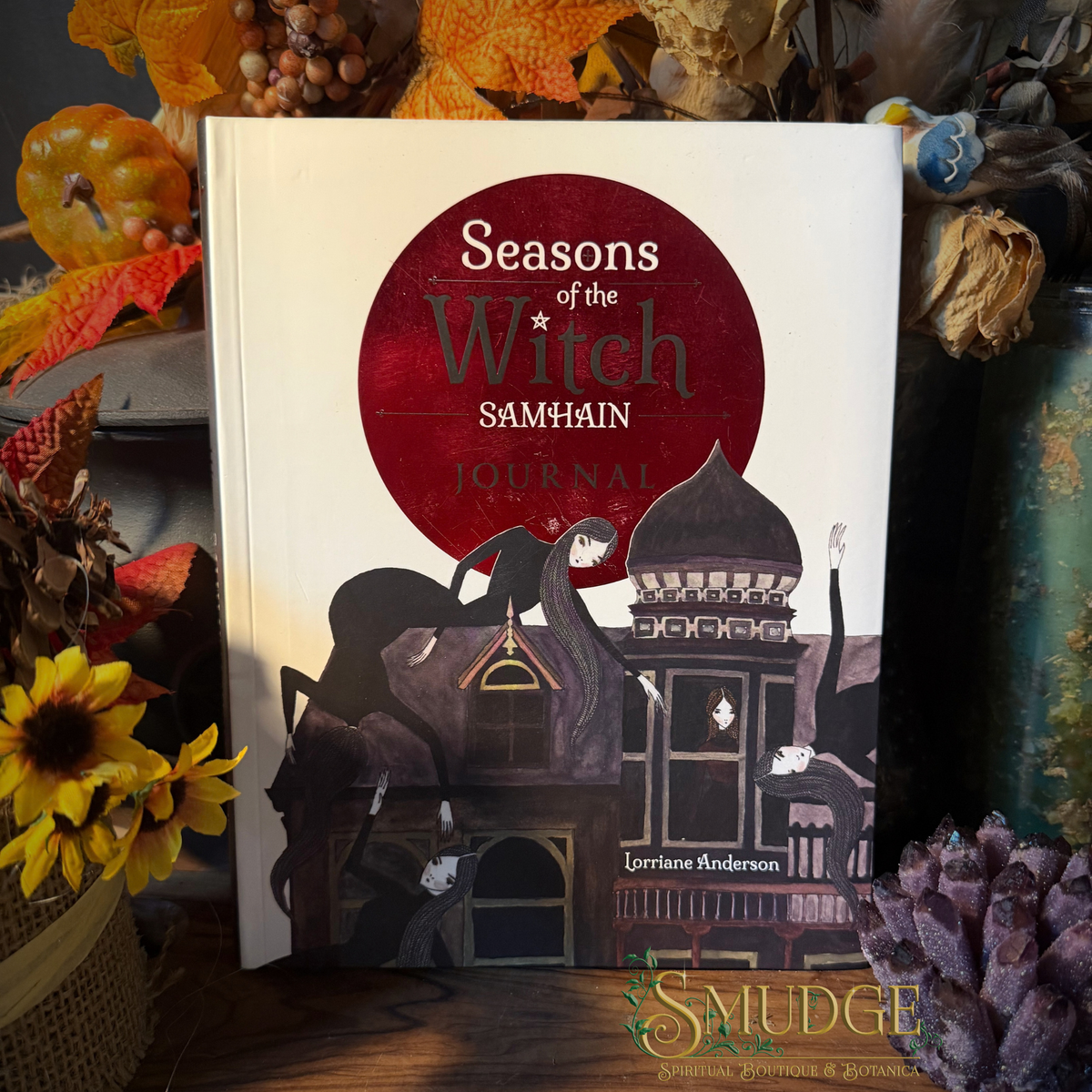A hardcover journal titled 'Seasons of the Witch Samhain' by Lorianne Anderson, displayed upright in a styled autumn setting with pumpkins, dried flowers, and a 'Smudge' brand watermark at the bottom.