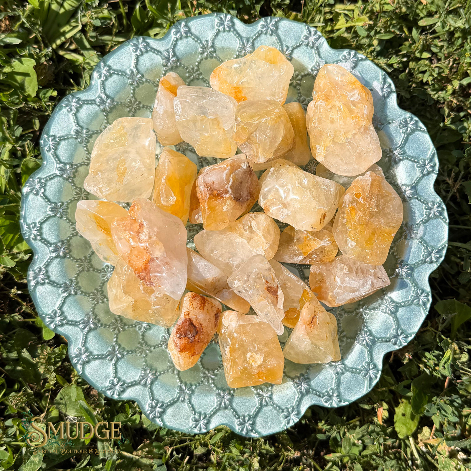 Raw crystal stones in pale yellow and orange tones arranged on a scalloped light blue dish placed outdoors on green grass. The stones appear translucent with natural rough edges.