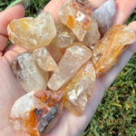 A hand holding a pile of rough, tumbled gemstone chips in natural outdoor light, with green grass visible in the background. The stones are translucent and have warm amber, orange, and cream tones.