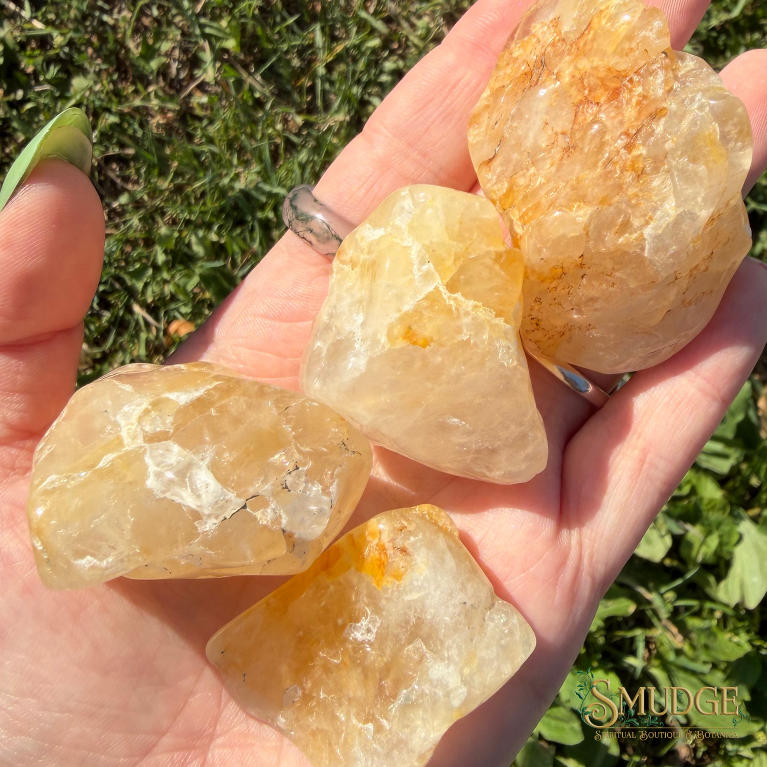 Four rough, unpolished yellow quartz crystal stones held in a person's hand outdoors on a grassy background. The stones have natural, jagged textures and a warm golden-yellow hue with some orange-brown mineral inclusions.