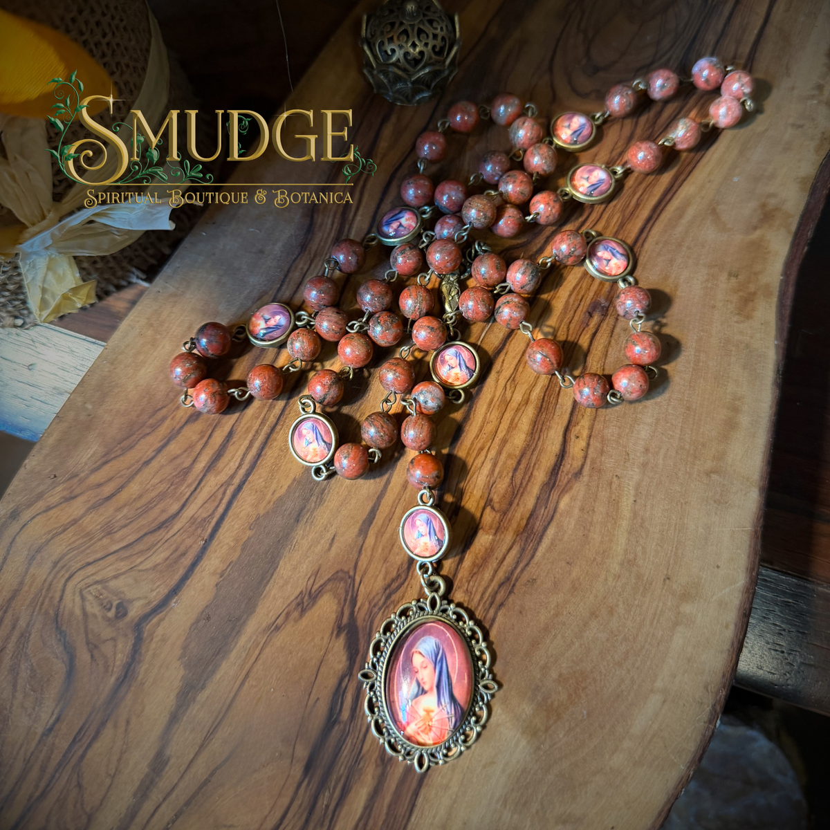 A religious rosary necklace with reddish-brown stone beads and ornate bronze-toned settings, laid out on a wooden surface. The centerpiece features a large oval medallion with a religious image, and several smaller oval links are visible along the chain.