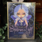 Oracle of Shadows & Light card deck box by Lucy Cavendish, featuring artwork by Jasmine Becket Griffith. The box is wrapped in protective plastic and displayed on a dark surface with green leaves in the background.