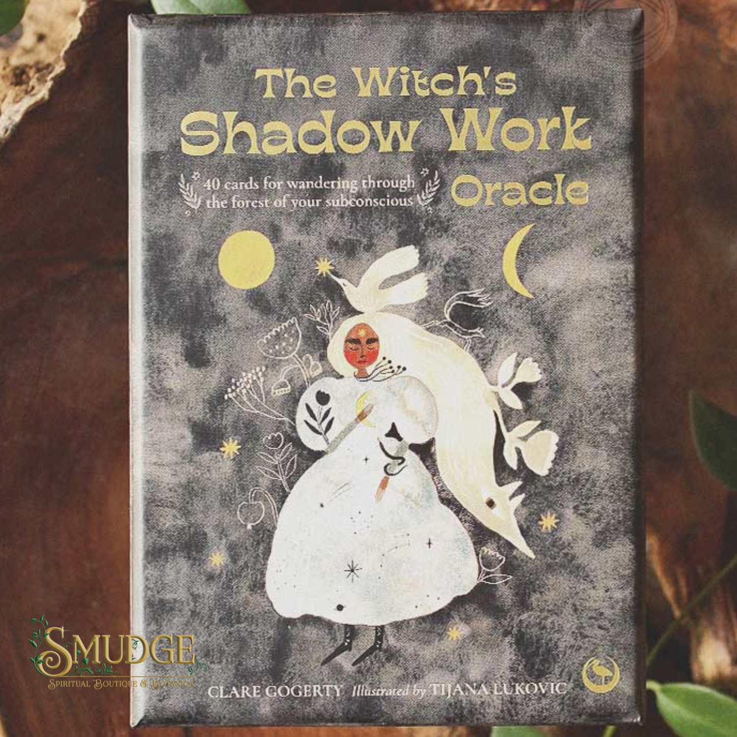 The Witch's Shadow Work Oracle card deck box by Smudge, featuring a dark textured cover with a white rabbit, yellow moon and sun, and gold lettering, photographed on a rustic wood surface.