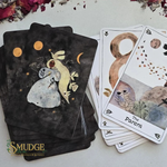 A deck of illustrated tarot or oracle cards from the brand Mudge, shown with a dark back design featuring a rabbit and celestial motifs, and several front cards fanned out including 'The Parent' card. The cards are arranged on a light surface with dried flower petals around the edges.