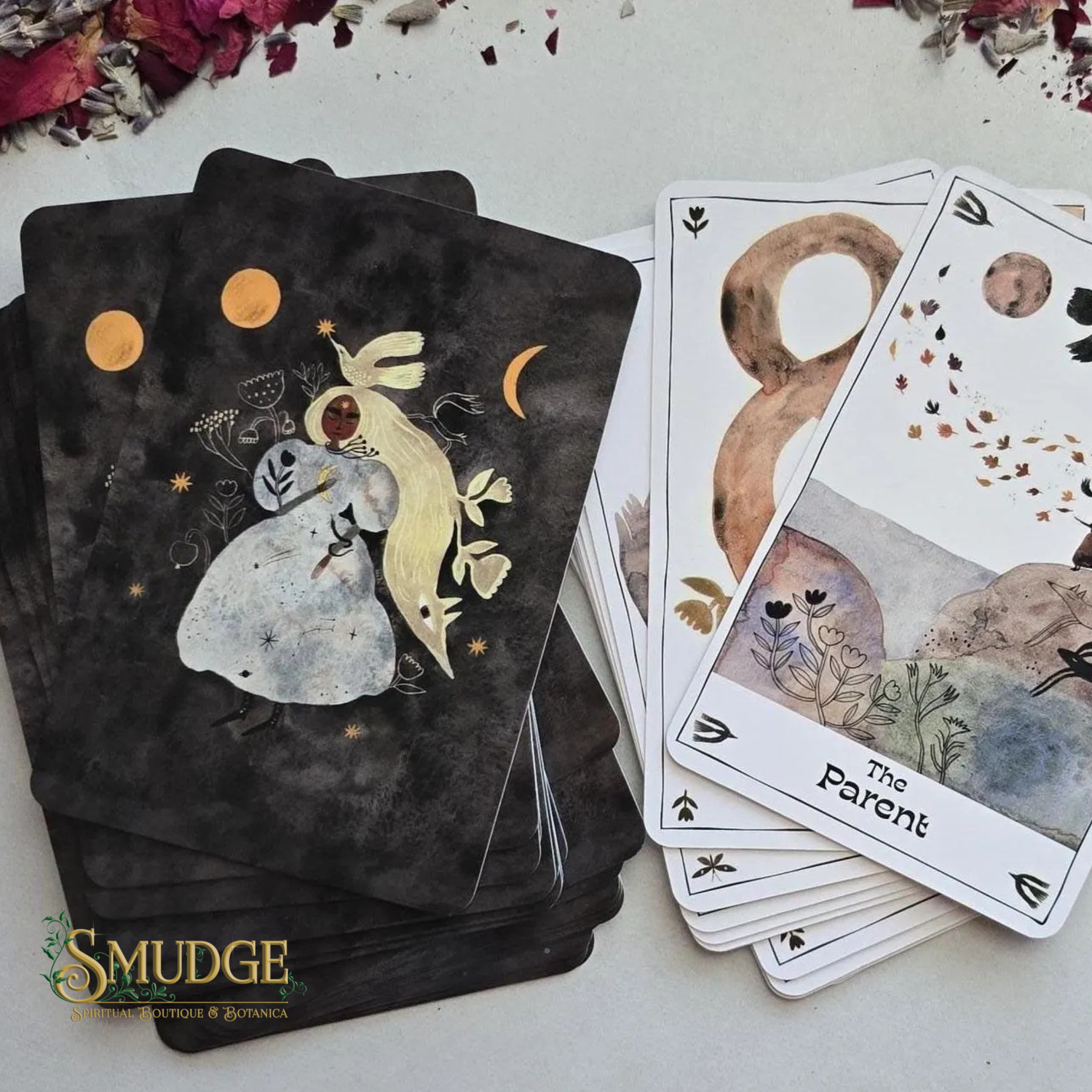 A deck of illustrated tarot or oracle cards from the brand Mudge, shown with a dark back design featuring a rabbit and celestial motifs, and several front cards fanned out including 'The Parent' card. The cards are arranged on a light surface with dried flower petals around the edges.