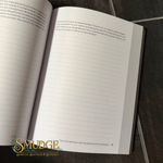 An open lined journal or notebook lying on a dark textured surface, with printed text and a visible 'SMUDGE Spiritual Boutique & Botanica' logo at the bottom left.