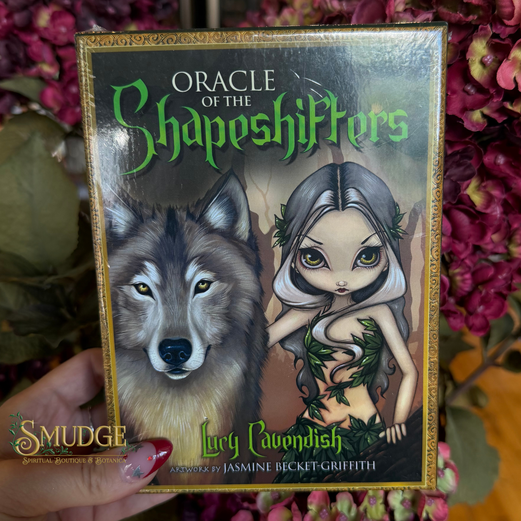 A hand holds the box of 'Oracle of the Shapeshifters' card game, featuring artwork of a wolf and a female figure with green vines, set against a background of pink flowers.