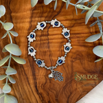 A silver-tone hamsa hand bracelet with blue and black eye details, displayed on a wooden surface with eucalyptus leaves. The brand logo 'SMUDGE' is visible in the bottom right corner.