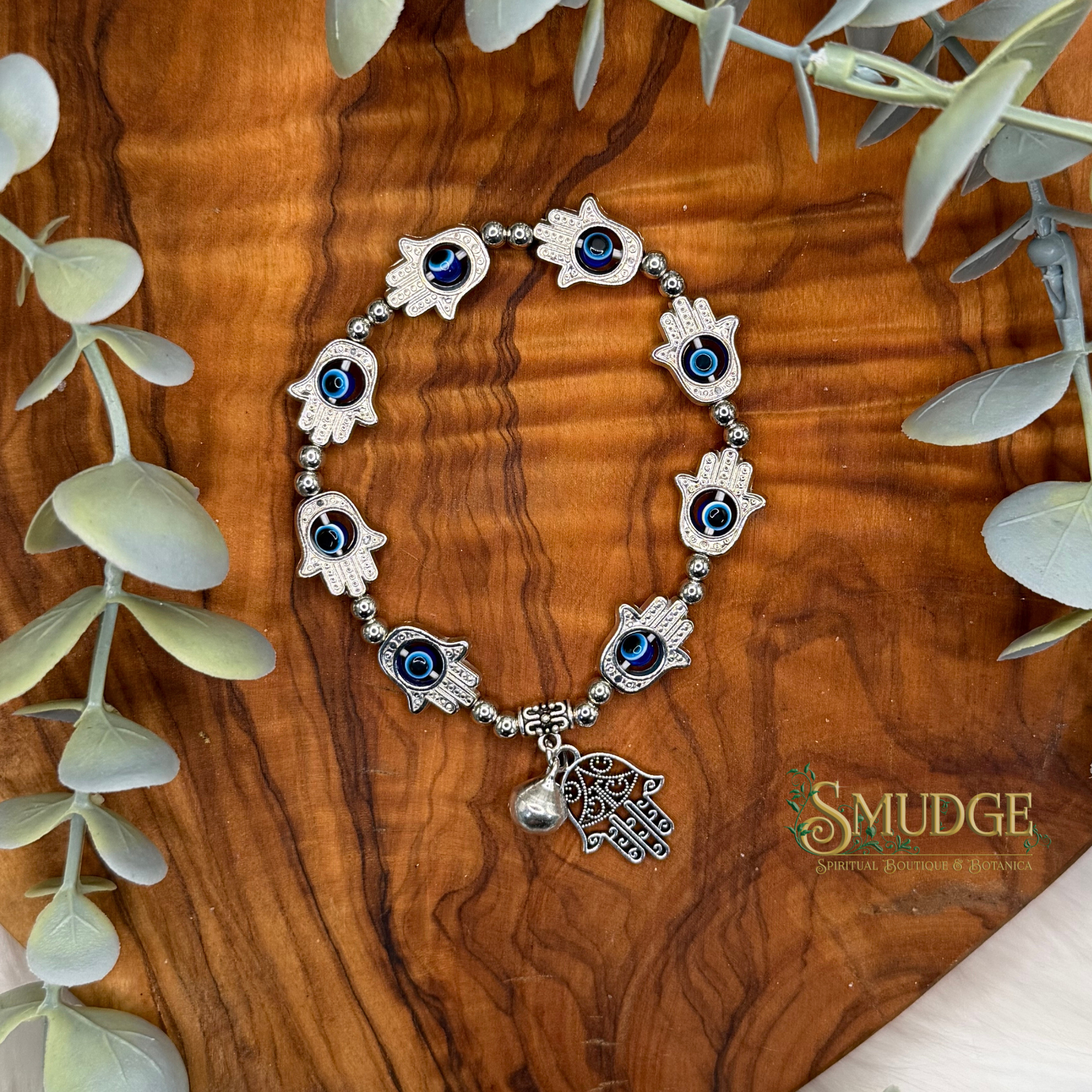 A silver-tone hamsa hand bracelet with blue and black eye details, displayed on a wooden surface with eucalyptus leaves. The brand logo 'SMUDGE' is visible in the bottom right corner.