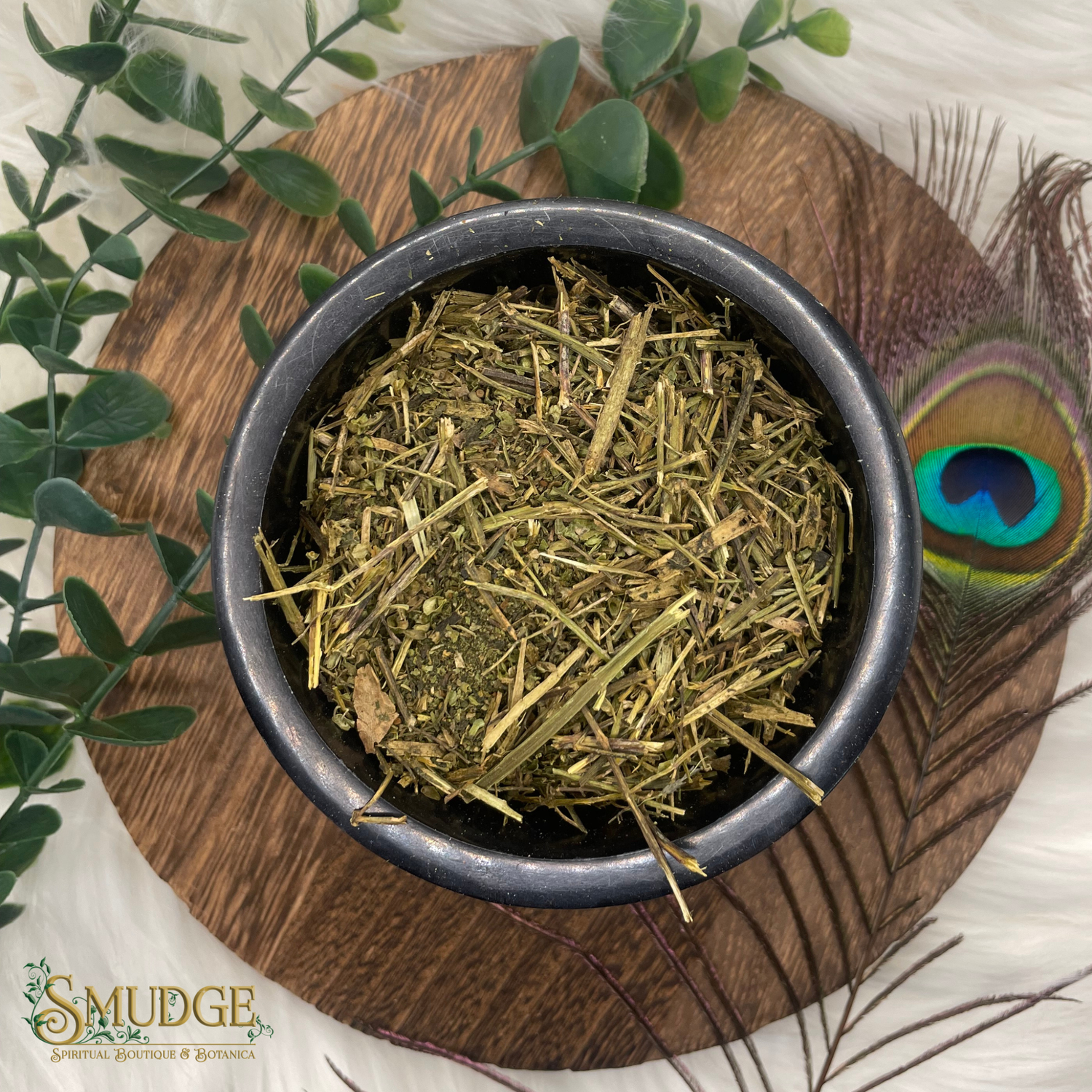 Dried herbs in a black bowl on a wooden coaster with a peacock feather and green leaves.