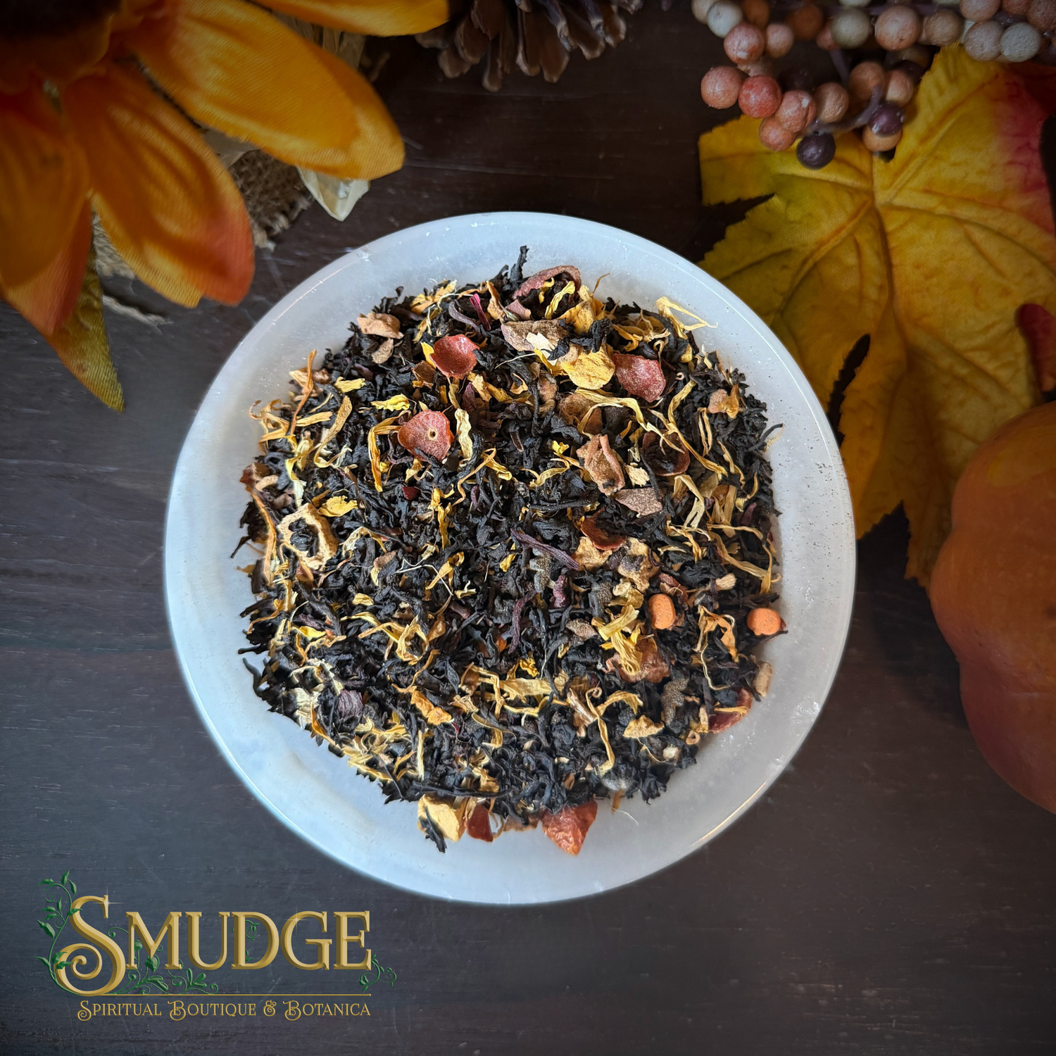 A top-down view of a white bowl filled with a loose-leaf herbal tea blend from Smudge Spiritual Boutique & Botanica, surrounded by autumn-themed decorative leaves, berries, and a pumpkin on a dark wooden surface.