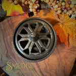 A round cast iron diffuser with a decorative starburst cutout pattern and a matching lid, displayed on a wooden surface with autumn leaves and dried grapes. The brand name 'SMUDGE' is visible in the bottom-left corner.