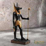 An Egyptian Anubis statue with a black body and gold details, holding a gold staff, standing on a rectangular base against a textured stone wall background.