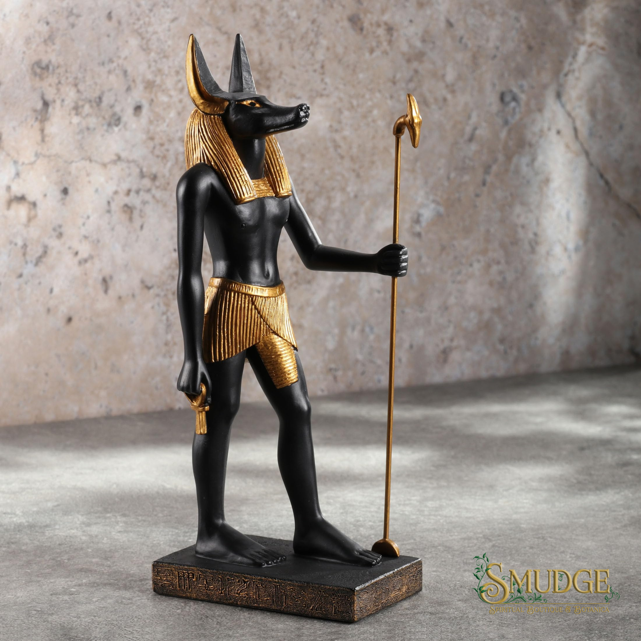 An Egyptian Anubis statue with a black body and gold details, holding a gold staff, standing on a rectangular base against a textured stone wall background.