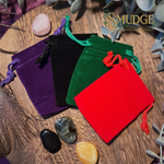 Four velvet bags in purple, black, green, and red on a wooden surface with crystals.