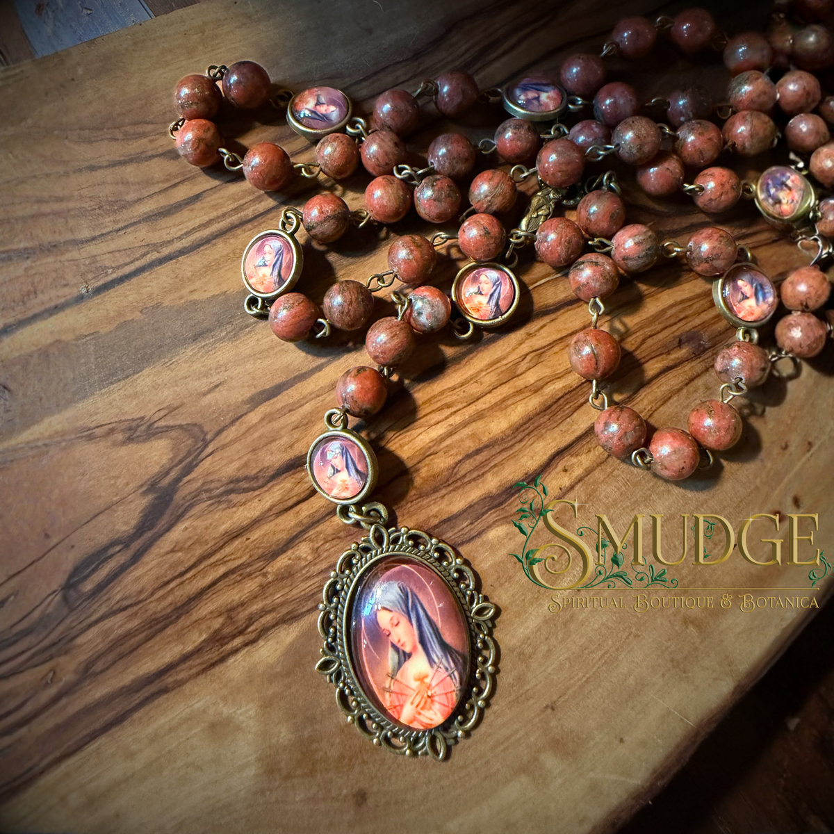 A religious rosary necklace with reddish-brown wooden beads, ornate bronze-toned metal settings, and oval medallions featuring a Virgin Mary, displayed on a rustic wooden surface. The brand name 'SMUDGE' is visible as a watermark overlay.