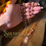 A hand holds a beaded rosary with reddish-brown stone-like beads and a detailed religious medallion pendant, displayed over a wooden surface. The brand name 'SMUDGE Spiritual Boutique & Botanicals' is visible as an overlay.