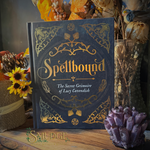 A hardcover book titled "Spellbound: The Secret Grimoire of Lucy Cavendish" stands upright on a wooden surface, surrounded by autumn-themed decorative props including dried flowers, sunflowers, and a purple crystal cluster. The cover is dark blue with ornate gold illustration and lettering.